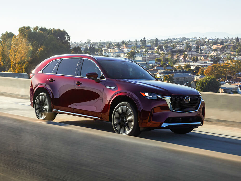 Un CX-90 rouge artisan roule sur un pont autoroutier en béton reflétant la lumière du jour.   