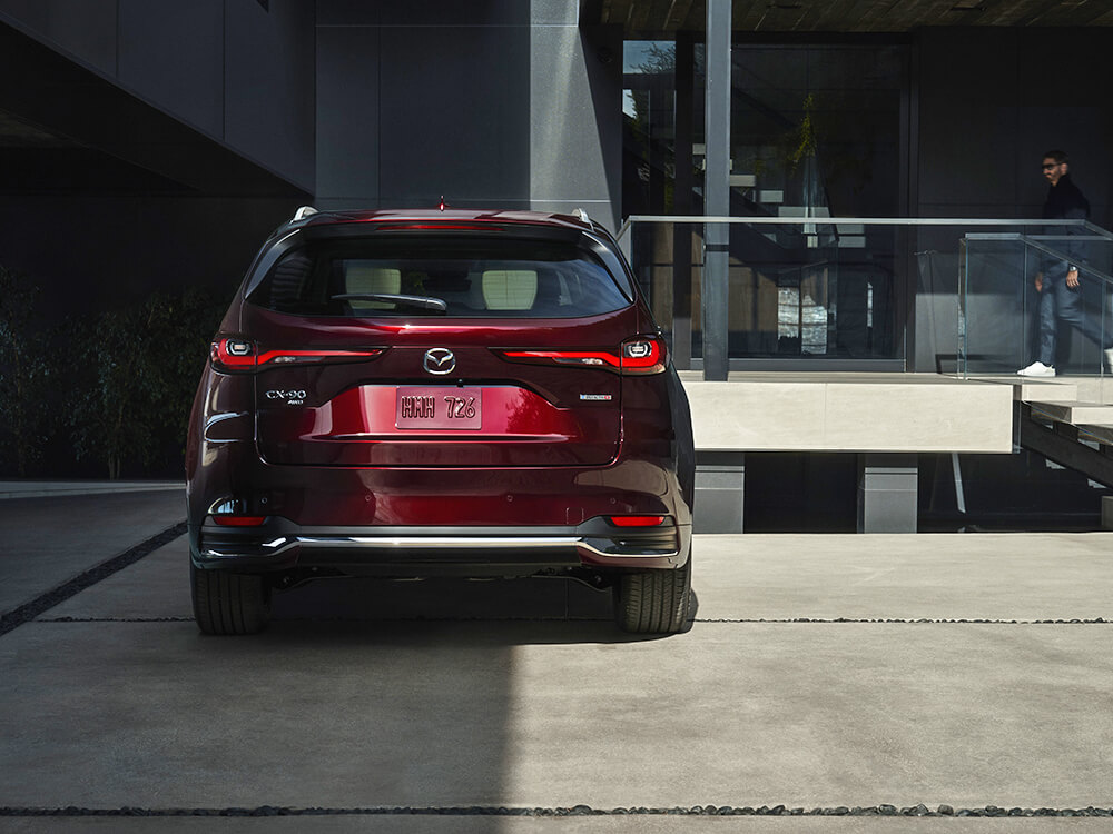 L’arrière d’un CX-90 rouge artisan garé devant l’atrium d’un bâtiment en béton avec des garde-corps en verre. 