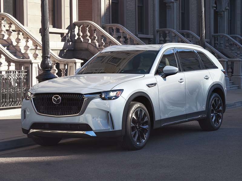 Un CX-90 blanc arctique garé dans une rue urbaine bordée d’escaliers d’appartements en béton.  