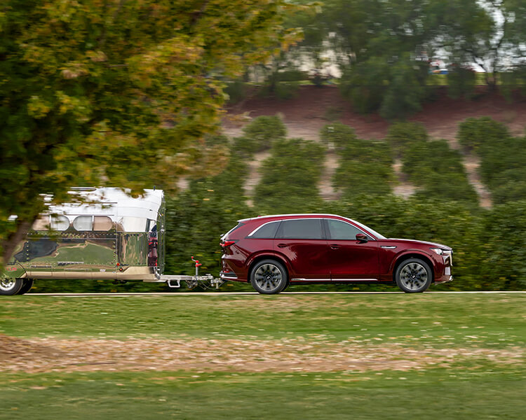 Un VÉHR CX-90 rouge artisan tracte une remorque de camping sur une route de campagne verdoyante. 