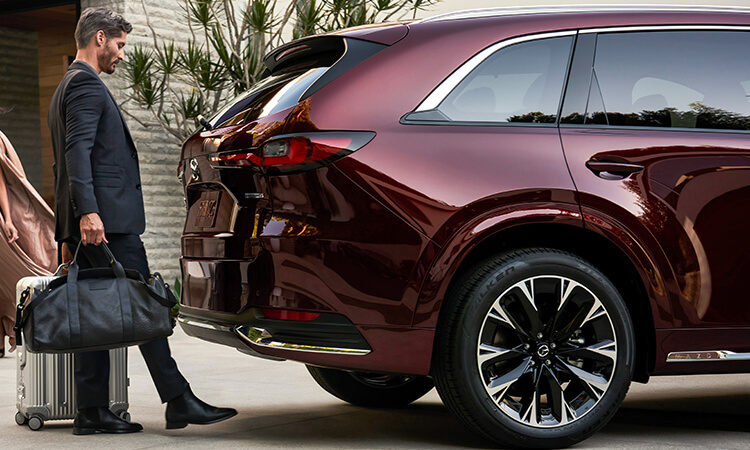 Un homme aux mains pleines utilise son pied pour activer le hayon électrique mains libres d’un CX-90 rouge artisan. 